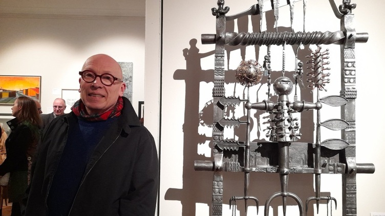 A person with glasses stands smiling beside an intricate metal sculpture displayed in an art gallery. The sculpture features various geometric shapes and patterns. Other visitors are visible in the background.