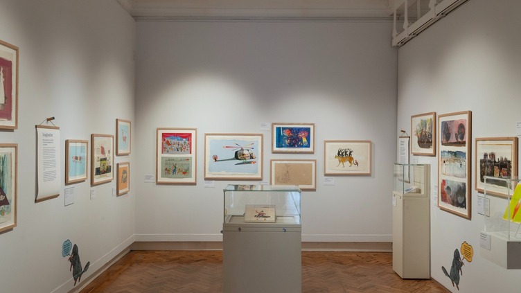 An art gallery room with framed artworks on white walls. A glass display case is in the center on a wooden parquet floor. The ceiling has track lighting and upper railings, and various illustrations are mounted around the space.